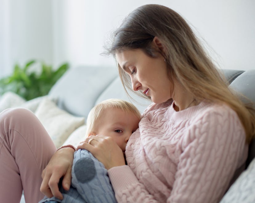 Young mother, holding her sick toddler boy, breastfeeding him at home, sunny living room