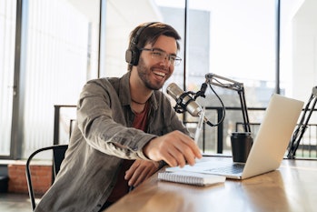 Young man host in headphones and glasses enjoying podcasting in studio, speaking into a microphone, ...