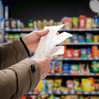 A person in a brown puffy coat looks at their receipts in the supermarket with a shelf of goods out ...