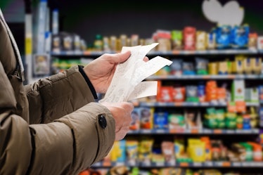 A person in a brown puffy coat looks at their receipts in the supermarket with a shelf of goods out ...