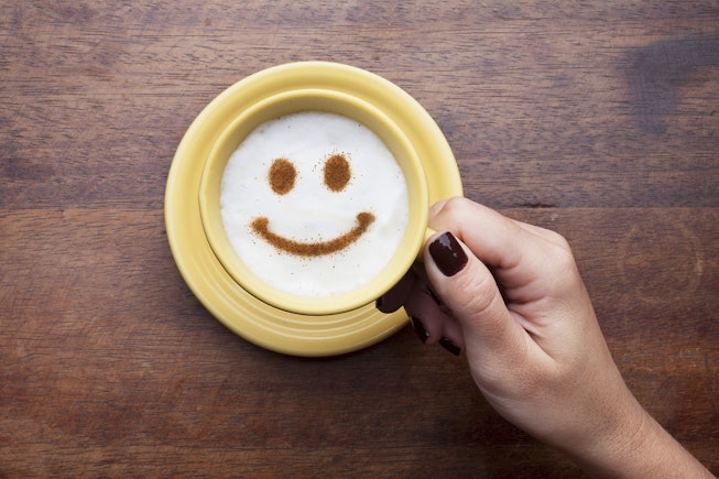 Hand holding a cute yellow cup with coffee cream. Food art creative concept image, happy face drawin...
