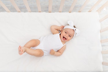 baby girl 6 months old lies in a crib in the nursery with white clothes on her back and laughs, look...