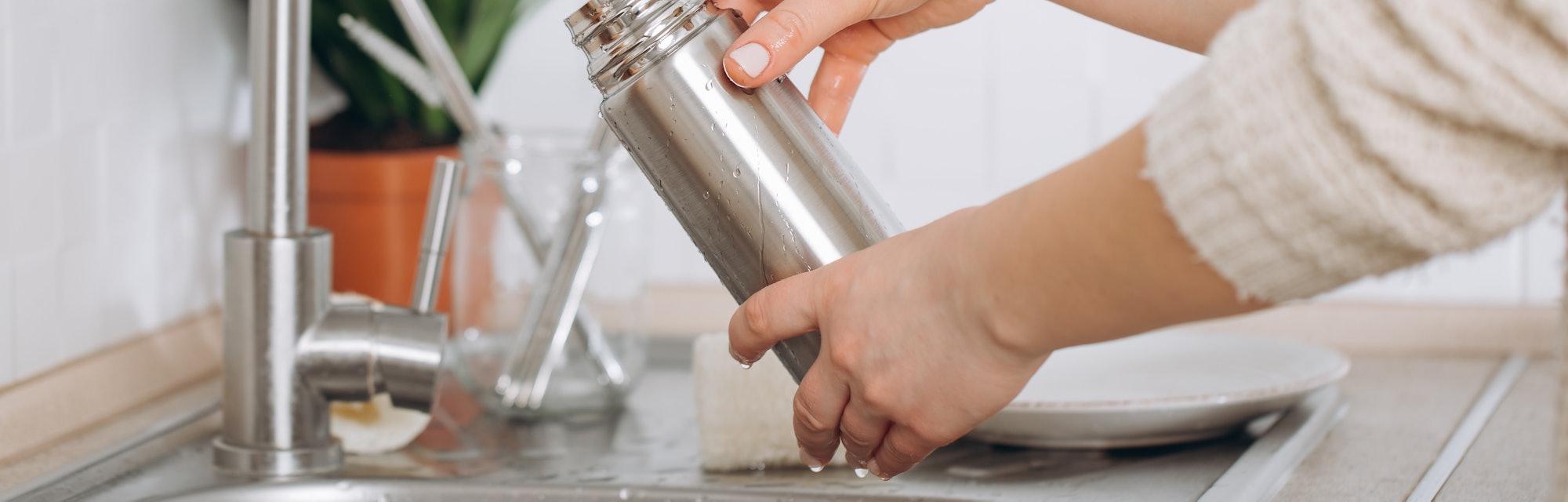 Two hands washing a metal reusable bottle in a sink, demonstrating how to clean a reusable water bot...