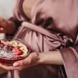 Unrecognizable pregnant female in silk robe eating porridge with strawberry and banana in morning at...