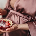 Unrecognizable pregnant female in silk robe eating porridge with strawberry and banana in morning at...