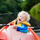 Happy kid enjoying kayak ride on beautiful river. Little curly toddler boy kayaking on hot summer da...