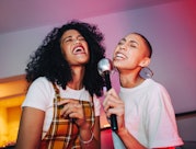 Best friends singing into a microphone on karaoke night. Two cheerful young women singing their favo...