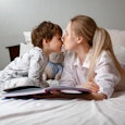 Cute preschool child son holding a book reading fairy tales for mom, lying in bed together, happy mo...