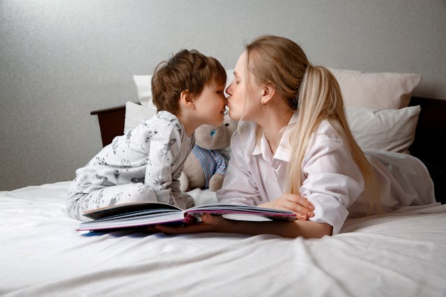 Cute preschool child son holding a book reading fairy tales for mom, lying in bed together, happy mo...