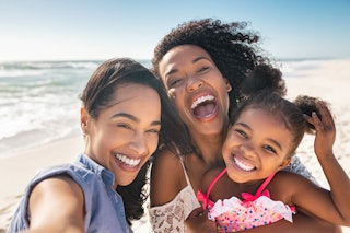 Portrait of smiling young african american woman with child taking selfie at beach with her best fri...