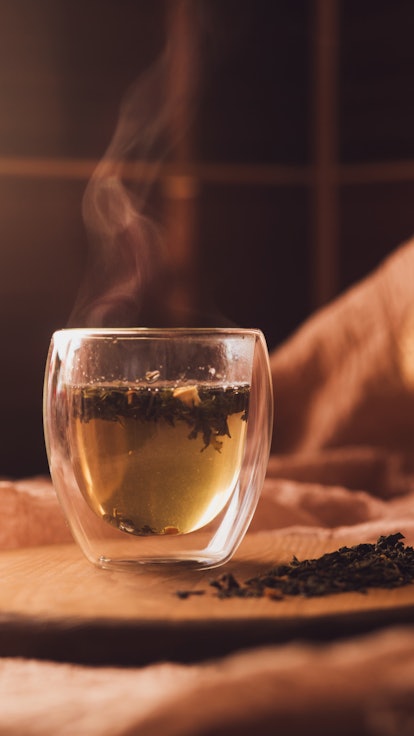 A glass cup of fragrant green tea on a wooden board next to scattered dry tea. steam over a hot drin...