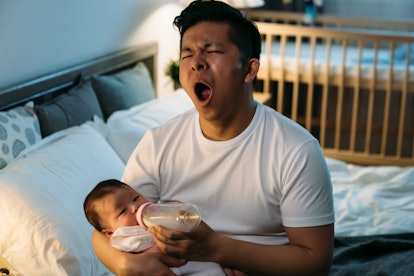 first time dad is yawning while sitting at bedside feeding his newborn girl for an article about how...