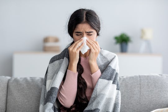 Woman blowing nose while she sits on a coucn under a blanket.