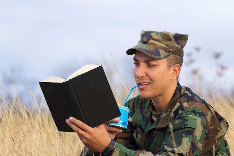 A soldier reads a book and drinks condensed milk from a tin can through a cocktail tube