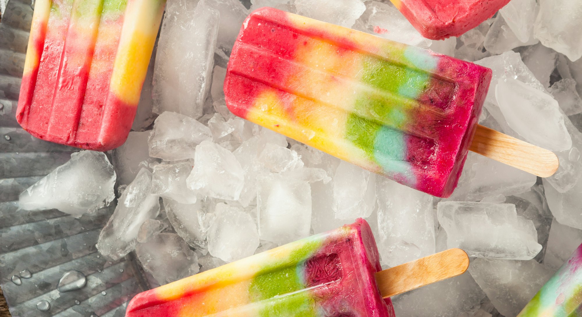 Homemade rainbow popsicles scattered on a tray of ice for summer popsicle inspiration.