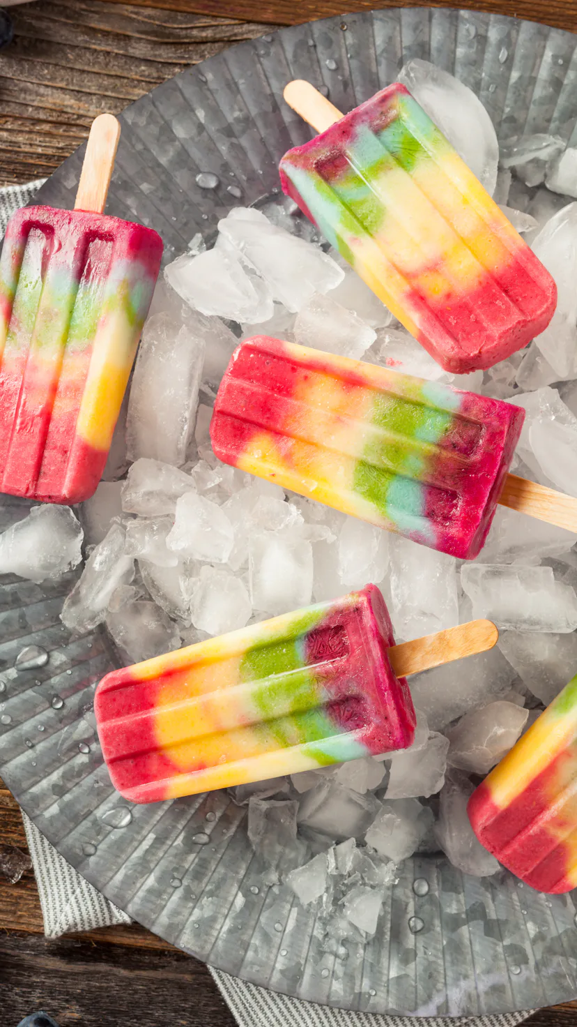 Homemade rainbow popsicles scattered on a tray of ice for summer popsicle inspiration.