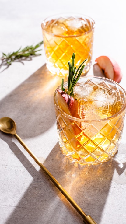 two ice cold bourbon peach smash cocktails with rosemary and stir spoon on white background with ros...