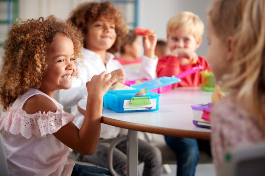 Close up of smiling young children sitting at a table eating their packed lunches together at infant...