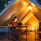 Cozy glamping tent with light inside and a woman using a laptop during dusk. Luxury camping tent for...