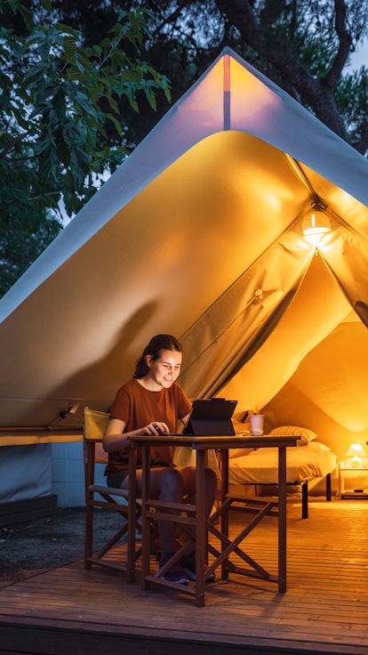 Cozy glamping tent with light inside and a woman using a laptop during dusk. Luxury camping tent for...