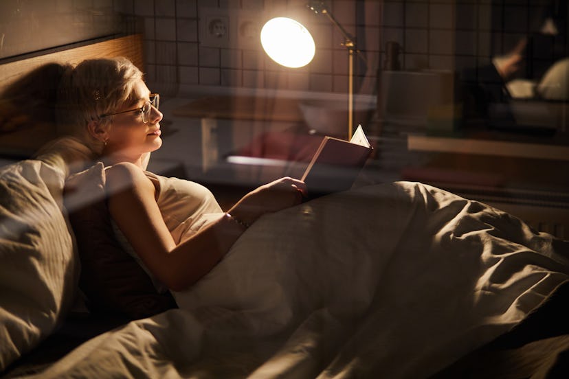 female in spectacles lying on bed reading favourite interesting book at home in room lighted by lamp