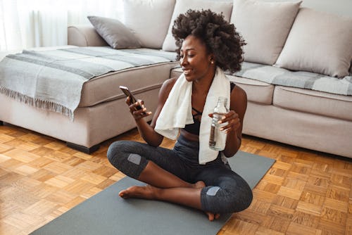 Woman using smart phone on exercise mat in front of her laptop. Sporty girl is exercising at phone o...