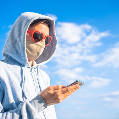 Woman wearing protective mask using mobile phone outdoors background. To shop online, texting messag...