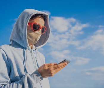 Woman wearing protective mask using mobile phone outdoors background. To shop online, texting messag...