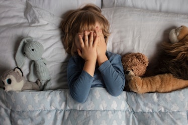 A kid lying in bed surrounded by stuffed animals and covering their face