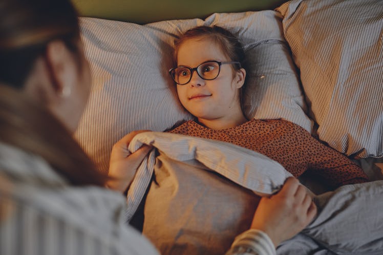 High angle portrait of caring mother tucking in child with down syndrome at bedtime in cozy lamp lig...