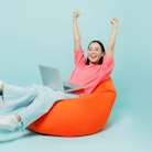 Full body young happy woman of Asian ethnicity 20s in pink sweater sit in bag chair use work on lapt...