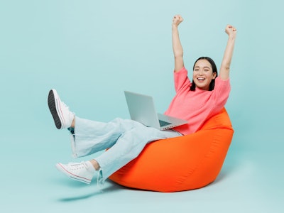 Full body young happy woman of Asian ethnicity 20s in pink sweater sit in bag chair use work on lapt...