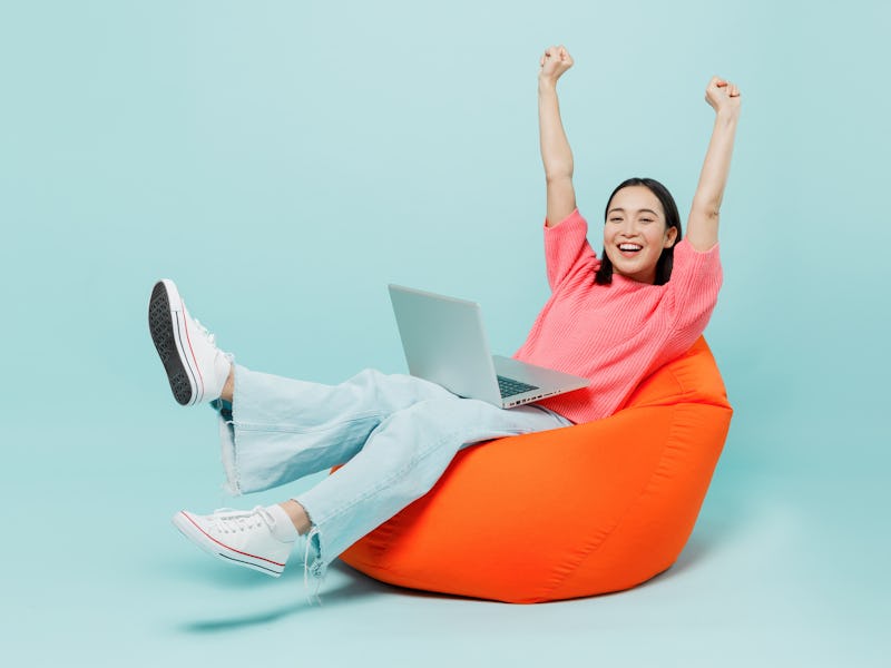 Full body young happy woman of Asian ethnicity 20s in pink sweater sit in bag chair use work on lapt...