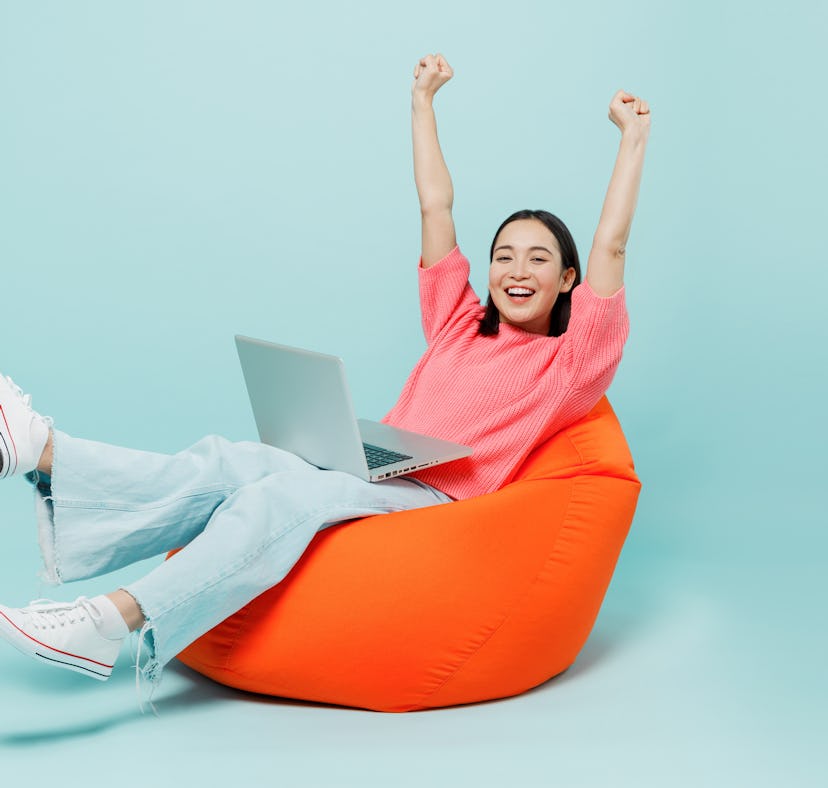 Full body young happy woman of Asian ethnicity 20s in pink sweater sit in bag chair use work on lapt...