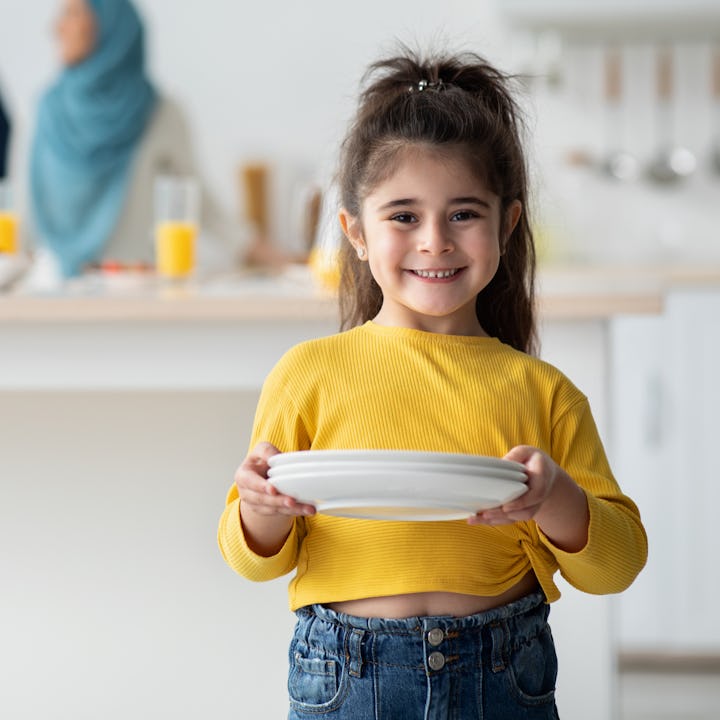 Little Helper. Portrait Of Cute Arab Girl Holding Plates In Kitchen And Smiling At Camera, Child Hel...