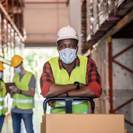 A Black male worker is wearing protective face mask and a bright yellow hazard vest while working in...