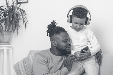 A Black dad and his son look at his son's phone, while his son wears blue headphones