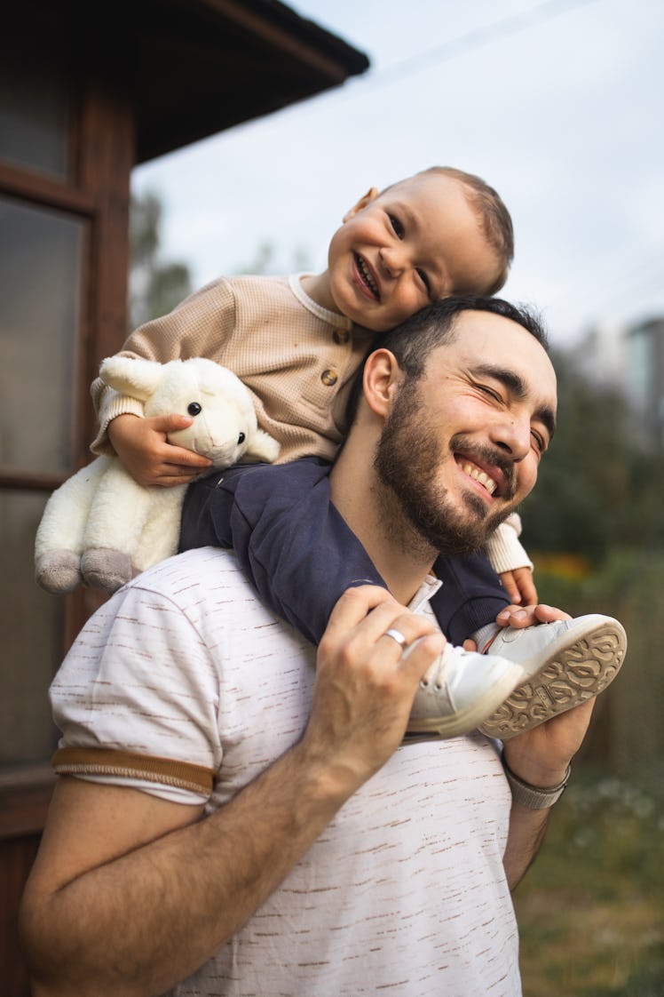 A dad stands with his son on his shoulders. The dad is smiling and so is the kid. The kid is holding...