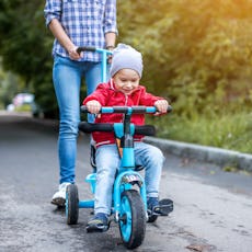 Young mother is pushing a child's tricycle with a toddler boy on a walk. Concept of learning to ride...