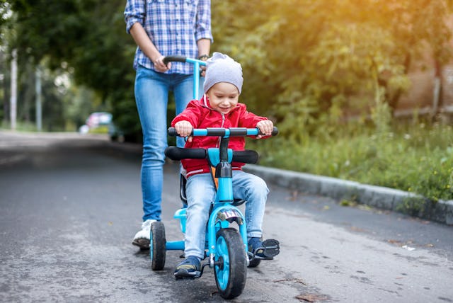 Young mother is pushing a child's tricycle with a toddler boy on a walk. Concept of learning to ride...