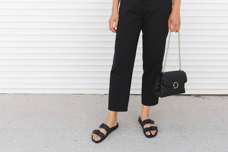 Bag in women's hands. Woman wearing black pants, bag with chain and flat sandals standing outdoor. D...