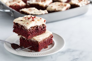 Two red velvet squares in a stack on a plate with other squares in soft focus in behind and copy spa...