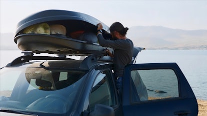 Young bearded man is opening roof bag on his crossover and taking out camping equipment. Traveler ca...