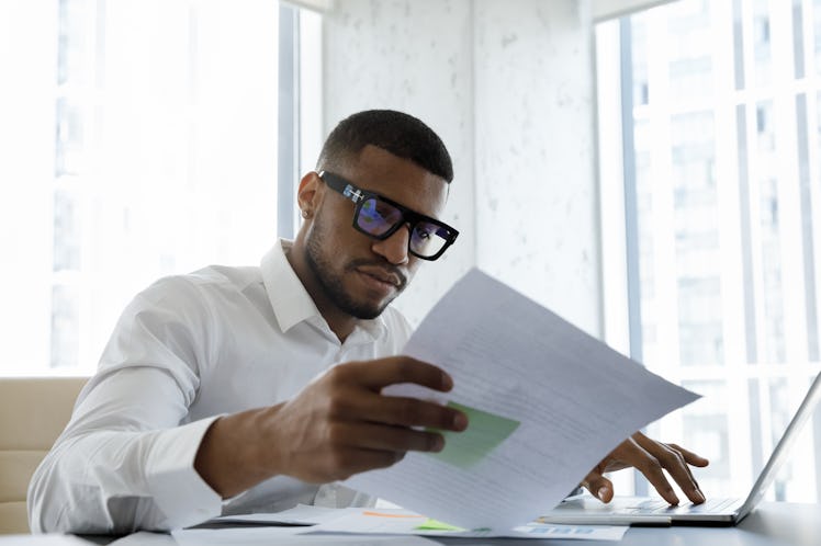 A Black man sits at his laptop, looking at a piece of paper. He wears glasses and a white, button up...