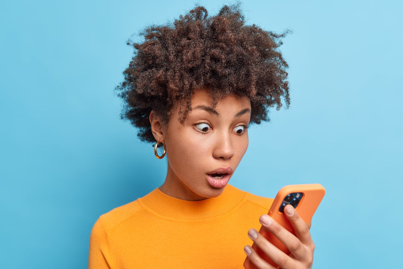 Emotional surprised African American woman stares at smartphone screen reads amazing news with surpr...