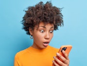 Emotional surprised African American woman stares at smartphone screen reads amazing news with surpr...