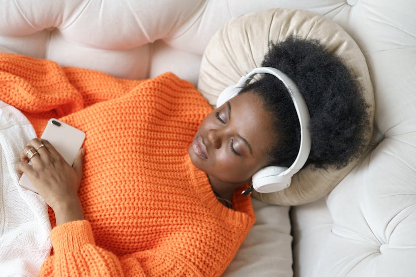 African millennial woman in orange sweater wearing wireless headphones, relaxes enjoying her favorit...