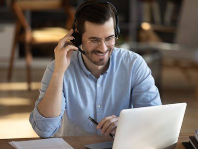 Smiling millennial Caucasian man in headset look at laptop screen work online from home office. Happ...
