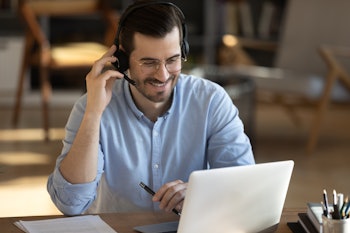 Smiling millennial Caucasian man in headset look at laptop screen work online from home office. Happ...