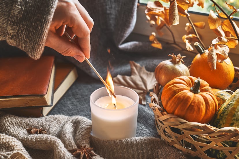 Hand with burning match lighting a candle next to cozy pumpkins in an article about candle instagram...